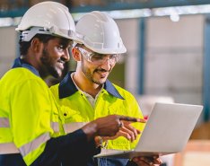 Two men in bright green work jackets, hard hats and safety gear looking at laptop