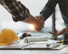 Two people shaking hands over a desk with hard hat and tape measure