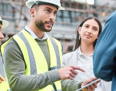 Workers in vests and hard hats gesturing toward phone and talking