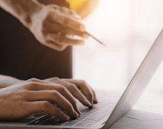 Hands on laptop while a person with hard hat and pencil gestures toward screen