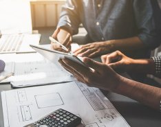 Two people sitting at desk, looking at the same tablet. Hard hat and blueprints on desk 